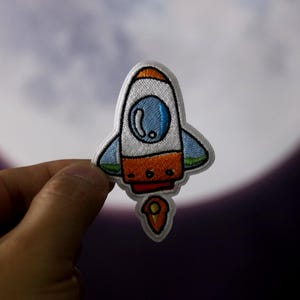 Könnte beinhalten: Ein gestickter Raketen-Patch in Weiß, Orange und Blau mit einer Flamme. Die Rakete hat ein rundes Fenster und eine kleine, spitze Nase.