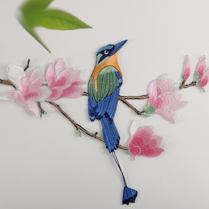 May include: Embroidered iron-on patch featuring a blue bird with a long tail perched on a branch with pink magnolia flowers.