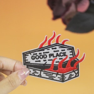 May include: A black and white embroidered patch of a coffin with flames coming out of it. The coffin has the words "GOOD PLACE" written on it.