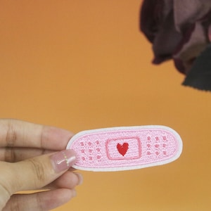 May include: A pink embroidered patch shaped like a bandage with a red heart in the center.