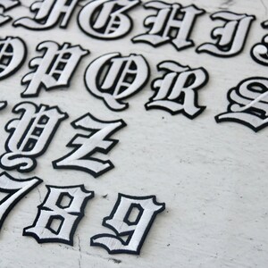 May include: White iron-on letters and numbers in a gothic font style. The letters are outlined in black and the interior is white. The letters include A through Z and the numbers 6 through 9.
