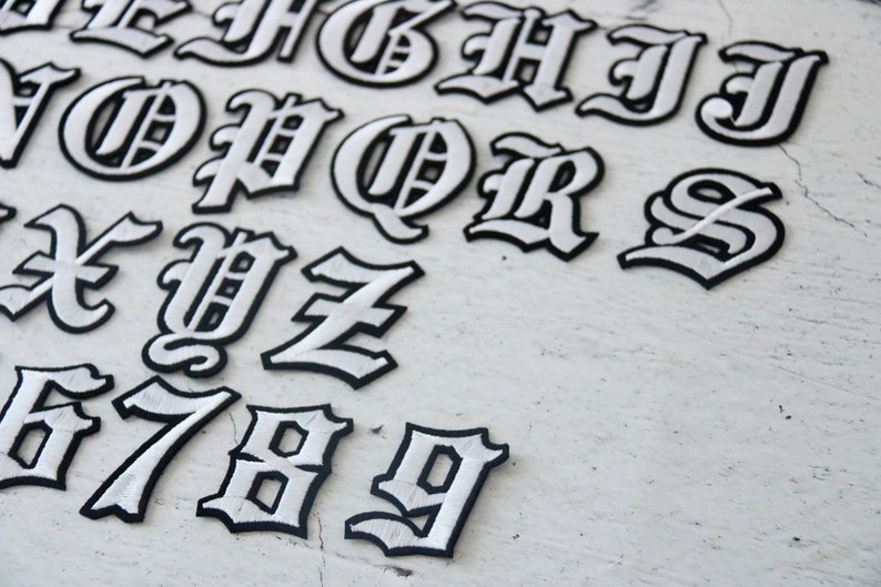 May include: White iron-on letters and numbers in a gothic font style. The letters are outlined in black and the interior is white. The letters include A through Z and the numbers 6 through 9.