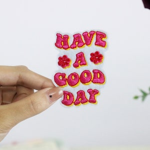 May include: A white patch with the text "HAVE A GOOD DAY" in pink letters with yellow outlines. The letters are in a retro font style with flowers on either side of the "A".