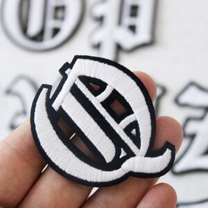 May include: White embroidered letter Q patch with a black outline. The patch is in a classic old English font style.