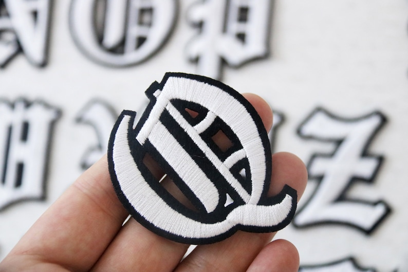 May include: White embroidered letter Q patch with a black outline. The patch is in a classic old English font style.
