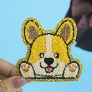 May include: A yellow and brown embroidered patch of a cartoon dog with a gold glitter outline. The dog is smiling and has its paws out.