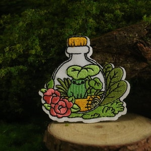 Może przedstawiać: Naszywkę haftowaną przedstawiającą szklaną butelkę z zieloną żabą w środku. Butelka jest wypełniona zielonymi liśćmi i różowym kwiatem. Naszywka jest otoczona zielonymi liśćmi.