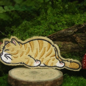 Puede incluir: Un parche bordado de un gato durmiendo, con pelaje marrón y naranja, patas blancas y cara blanca. El parche está sobre una superficie de madera con un hongo rojo y blanco a cada lado.