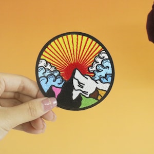 Może przedstawiać: Okrągła naszywka haftowana przedstawiająca kolorową scenę górską. Naszywka zawiera czerwone słońce z żółtymi promieniami, niebieskie chmury i zieloną górę z białym szczytem. Naszywka idealnie nadaje się do dodania odrobiny natury do ubrań lub akcesoriów.