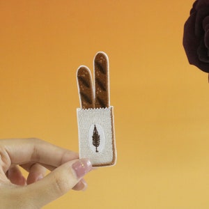 May include: A brown and white embroidered patch depicting a bag of bread. The bag is white with brown stitching and a small brown tree logo. The bread is brown and sticking out of the top of the bag.