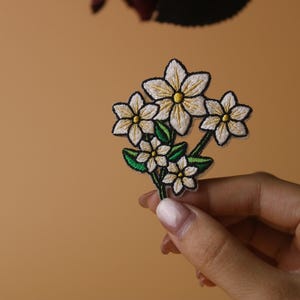 May include: A white embroidered flower patch with green leaves. The patch is held in a hand.