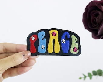 May include: A colourful embroidered patch with the word "PEACE" in a retro font. The letters are outlined in black and have white flowers embroidered on them.