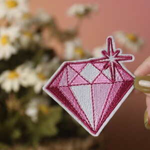 May include: Pink diamond shaped embroidered patch with a white starburst design. The patch is iron-on and can be used to decorate clothing, bags, or other items.