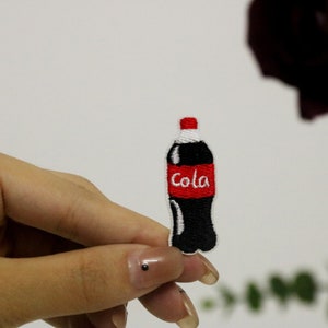 Op de afbeelding: Een geborduurde strijk-op patch van een cola-fles in zwart en rood met het woord "Cola" in witte letters.