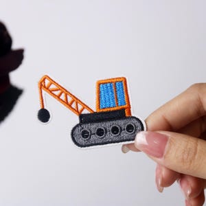 May include: An orange and blue embroidered iron-on patch of a crane with a black ball on the end of the arm.