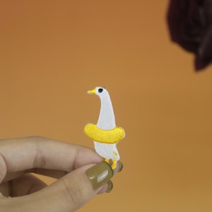 Puede incluir: Un parche bordado de un ganso con un salvavidas amarillo. El ganso mira hacia la derecha y tiene un ojo negro.