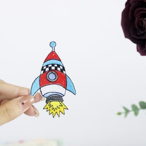 May include: A red, white and blue rocket ship with a chequered pattern on the side. The rocket has a yellow flame coming out of the back.