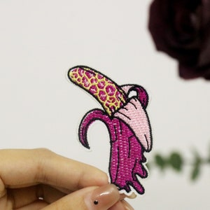Könnte beinhalten: Ein gestickter Bananen-Patch in Pink und Gelb mit einem Tropfeffekt. Die Banane ist geschält und das Innere ist rosa.