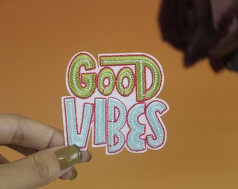 May include: A white embroidered patch with the words "Good Vibes" in green and blue lettering with red outlines.