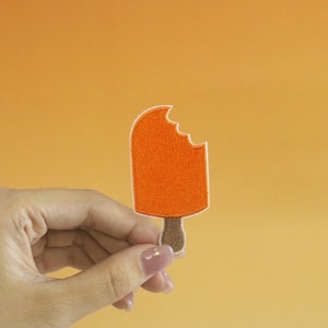 Pode incluir: Patch de picolé laranja com uma mordida. O patch é segurado em uma mão contra um fundo amarelo.