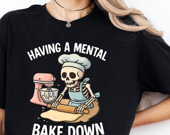 May include: Black t-shirt with the text "HAVING A MENTAL BAKE DOWN". The graphic features a skeleton chef with a chef's hat and apron, rolling dough with a rolling pin, and a pink stand mixer.