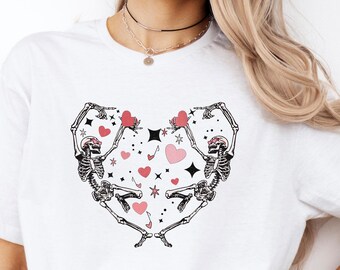 May include: White t-shirt with a graphic of two skeletons holding hands in the shape of a heart. The heart is filled with pink and red hearts, stars, and other small details.