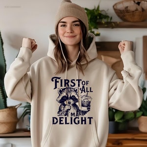 May include: A beige hoodie with a raccoon graphic holding a coffee cup and the text "First of all I'm a delight".