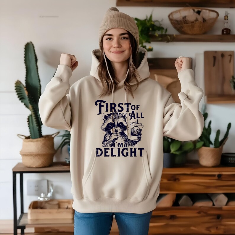 May include: A beige hoodie with a raccoon graphic holding a coffee cup and the text "First of all I'm a delight".
