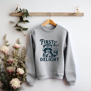 May include: A light gray sweatshirt with a graphic of a raccoon holding a cup of coffee. The text "First of all I make delight" is printed below the raccoon.