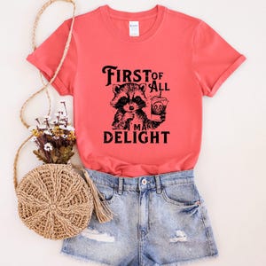 May include: A coral colored t-shirt with a black and white graphic of a raccoon holding a cup of coffee. The text on the shirt reads "First of all I'm a delight".
