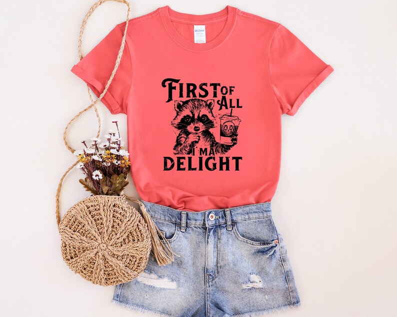 May include: A coral colored t-shirt with a black and white graphic of a raccoon holding a cup of coffee. The text on the shirt reads "First of all I'm a delight".