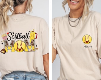 May include: A beige t-shirt with a graphic design on the back that says "Softball Mom" with a softball, popcorn, and a drink. The front of the shirt has a softball graphic and the name "Claire".