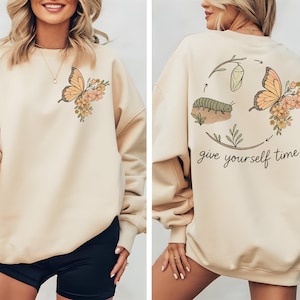 May include: Beige sweatshirt with a butterfly and floral design on the front and back. The back features a caterpillar, chrysalis, and butterfly with the text "give yourself time". The butterfly is orange and the flowers are yellow.