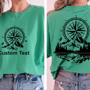May include: A green t-shirt with a black compass and mountain design. The text "Custom Text" is printed on the front of the shirt.