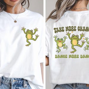 May include: White t-shirt featuring a cartoon frog on the front and back. The front has a single frog, while the back has three frogs with the text "TAKE MORE CHANCES, DANCE MORE DANCES."