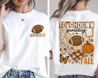 May include: White t-shirt with a football graphic and the word "TOUCHDOWN" on the front. The back features a "TOUCHDOWN gameday FALL" design with a football, pumpkin, and other fall-themed elements.