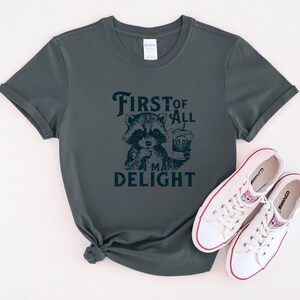May include: A dark green t-shirt with a raccoon graphic and the text "First of all I'm a delight".