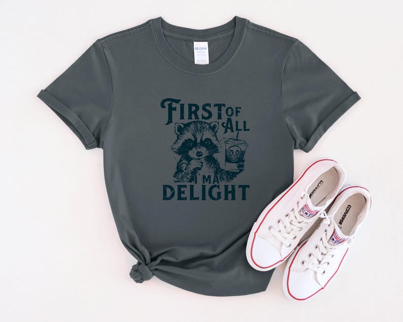 May include: A dark green t-shirt with a raccoon graphic and the text "First of all I'm a delight".