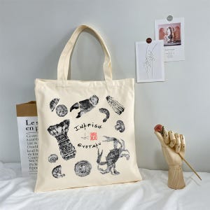 May include: Cream-colored tote bag featuring black ink illustrations of seafood, including a lobster tail and crab. The bag has the text "INKFISH GYOTAKU" and a red stamp. The bag has a handle.