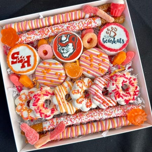 May include: A white gift box filled with a variety of treats, including pretzel rods dipped in white chocolate and sprinkles, chocolate-covered Oreos, and gummy candies. The treats are decorated with orange and pink sprinkles and designs. The box also includes a cupcake with pink frosting and a white chocolate cookie with the words "SHSU Bearkats" in blue.