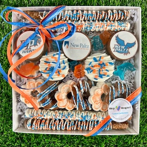 May include: A white gift box filled with a variety of treats, including chocolate-covered pretzels, chocolate-covered Oreos, and chocolate-covered strawberries. The box is decorated with blue and orange ribbon and a small tag that says "Hey Sugar! Custom Confections".