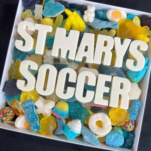 May include: A white box filled with assorted colorful candies. The candies include gummy bears, donuts, and other shapes in blue, yellow, and other colors. The words "ST MARYS SOCCER" are spelled out in white letters across the candies.