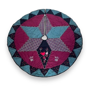 May include: A circular quilted tree skirt featuring a central star pattern in deep burgundy and teal plaid fabric. The design includes embroidered red bird and floral motifs, with a dark navy border of triangular points and stitched berry sprigs.