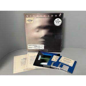May include: A vintage MINDSHADOW computer game box set. The box features a grayscale image of a face. Included are blue floppy disks, manuals, and promotional materials. The game is for Atari ST series computers.