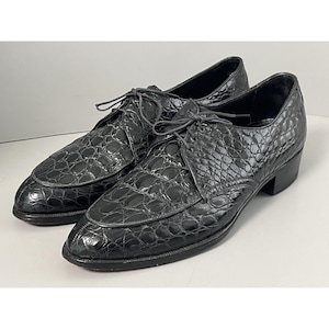 May include: A pair of black leather dress shoes with a textured, crocodile-like pattern. The shoes have a lace-up design and a low heel. The shoes are displayed on a white surface.