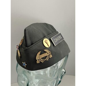 May include: Dark green military hat with gold-colored pins and a name tag that reads "CONVERSE." The hat features a gold torch pin, a gold ROTC emblem, and a blue star. A vintage military collectible.