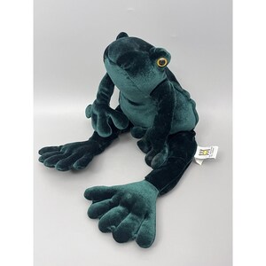 May include: A plush frog toy in a deep teal green color. The frog has large, expressive eyes with yellow irises. Its long arms and legs are outstretched, and it has a tag attached to one leg.