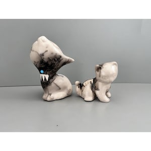May include: Two ceramic figurines. One is a stylized creature with a large head, black markings, and a blue eye. The other is a small cat with similar black markings. Both are on a gray surface.