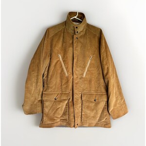May include: A tan corduroy jacket featuring a high collar with a snap button closure, a central zipper placket, two diagonal zippered chest pockets, and two large lower patch pockets with snap button flaps, all rendered in a textured fabric.