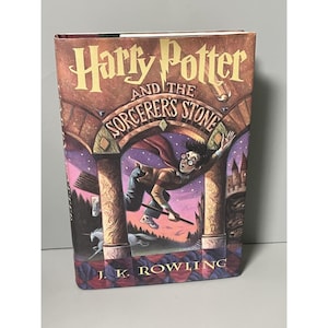 May include: A hardcover book titled Harry Potter and the Sorcerer's Stone by J.K. Rowling. The cover features a colorful illustration of a character flying on a broomstick through a stone archway, with a unicorn visible in the background.